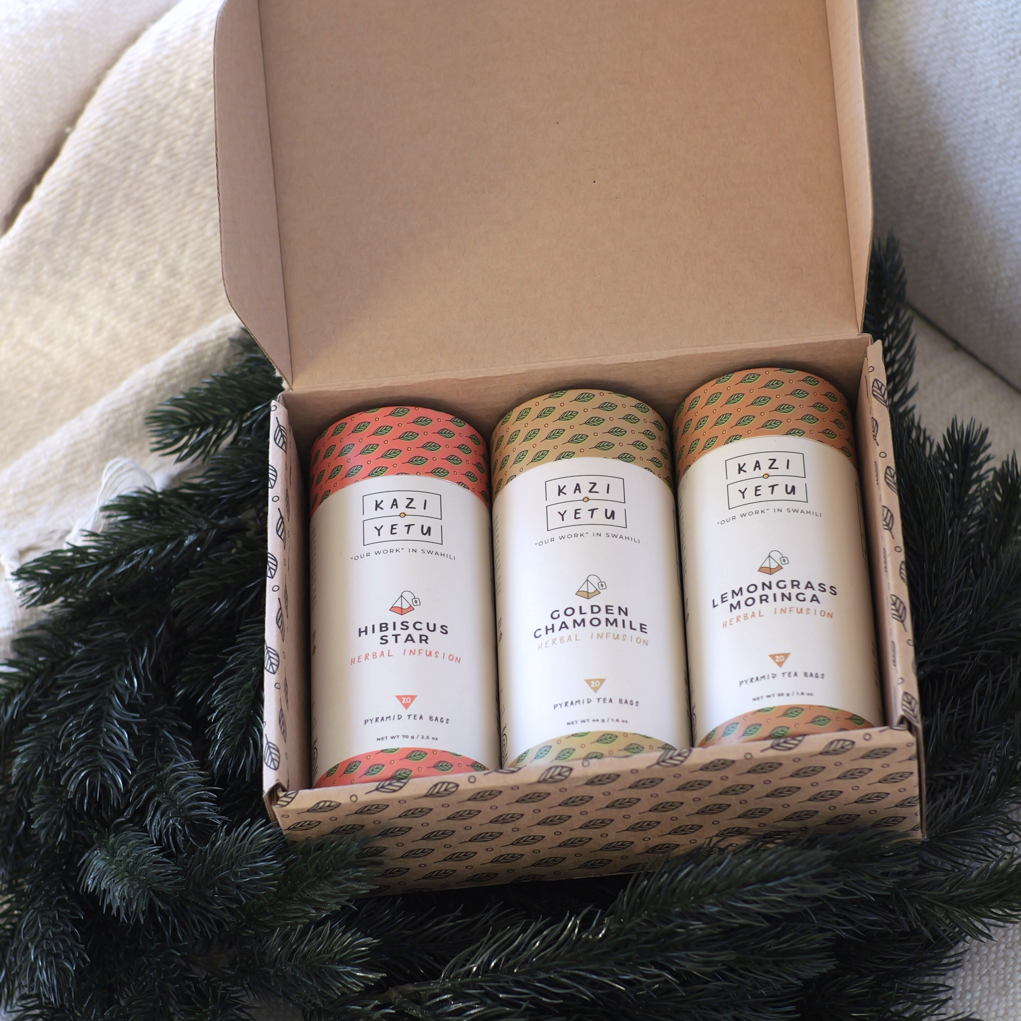 Three Kazi Yetu tea belnds in a cardboard box with decorative greenery around.