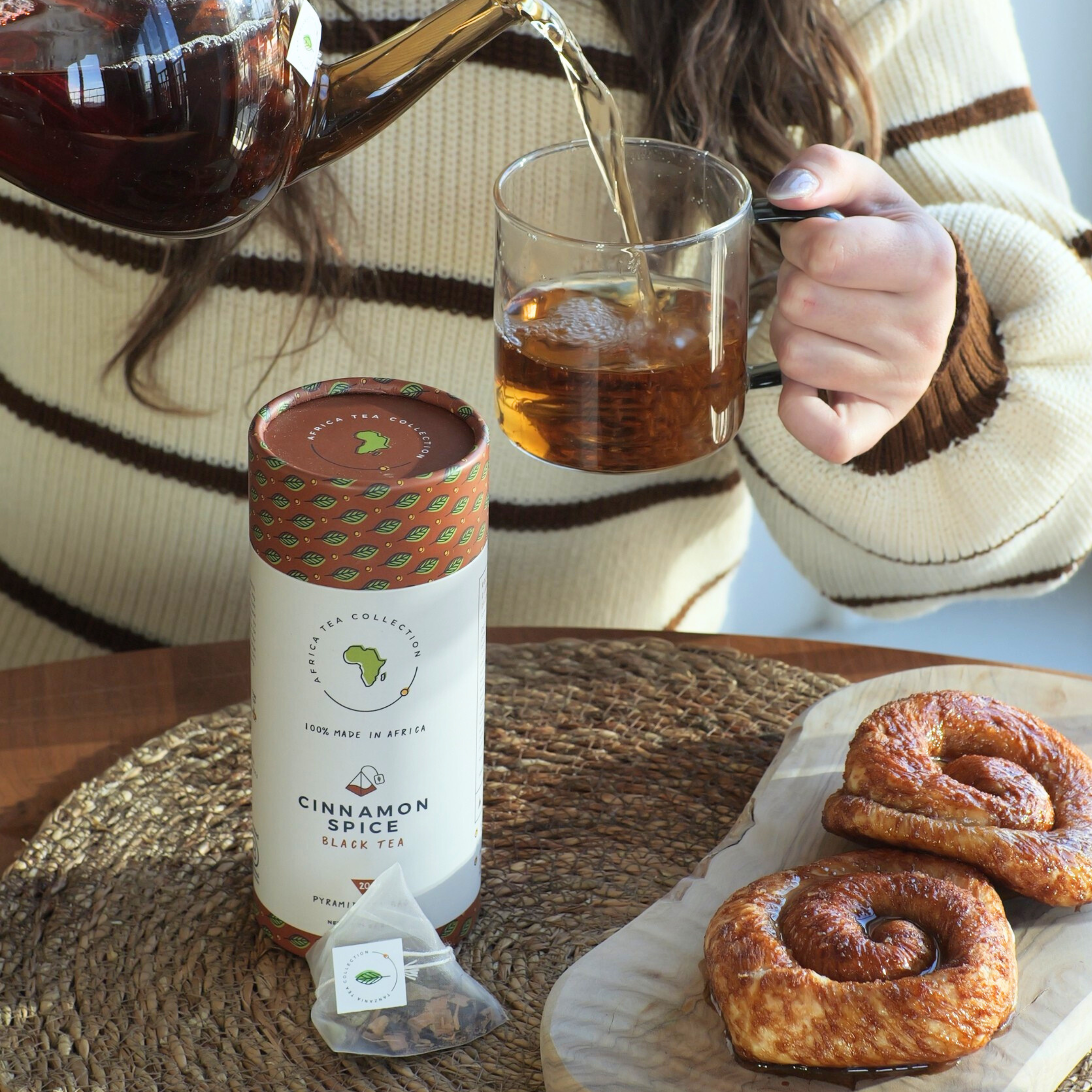 A woman is pouring Cinnamon Spice tea into a glass mug, with a package of Cinnamon Spice tea and pastries on the table.