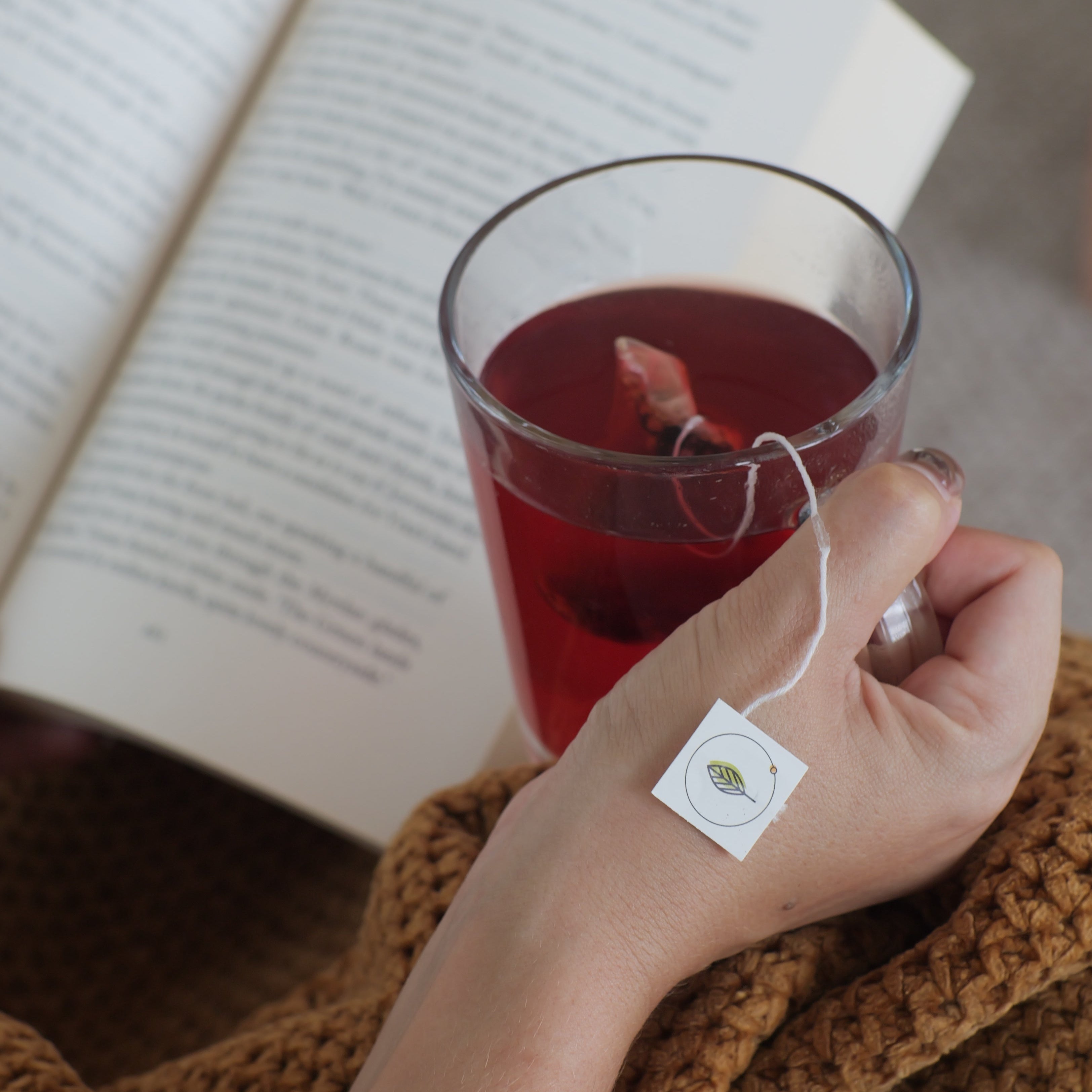Hand holding a glass of Hibiscus Star tea with a teabag, next to an open book.