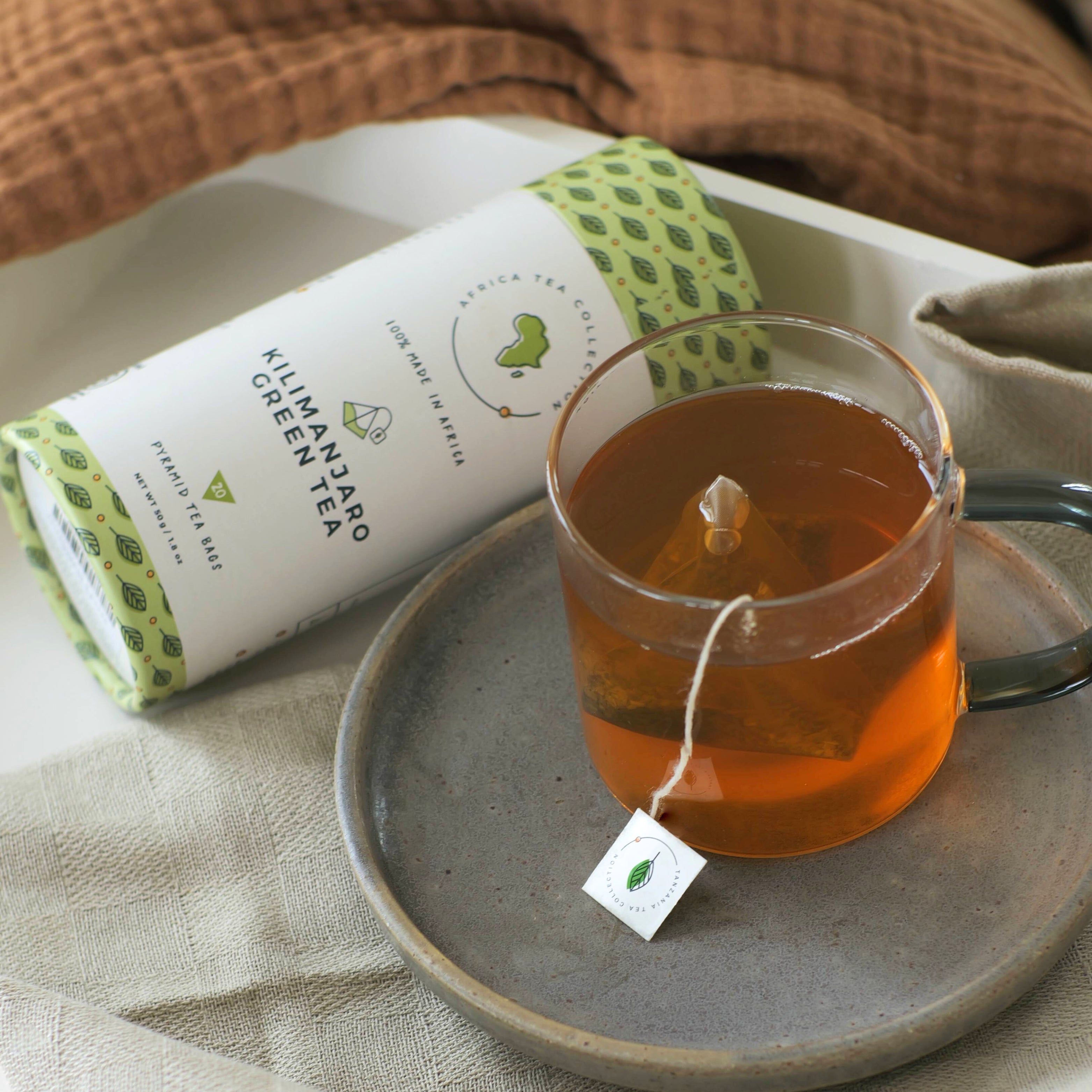 Glass mug of green tea with a tea bag on a gray plate, next to a box of Kilimanjaro Green Tea.